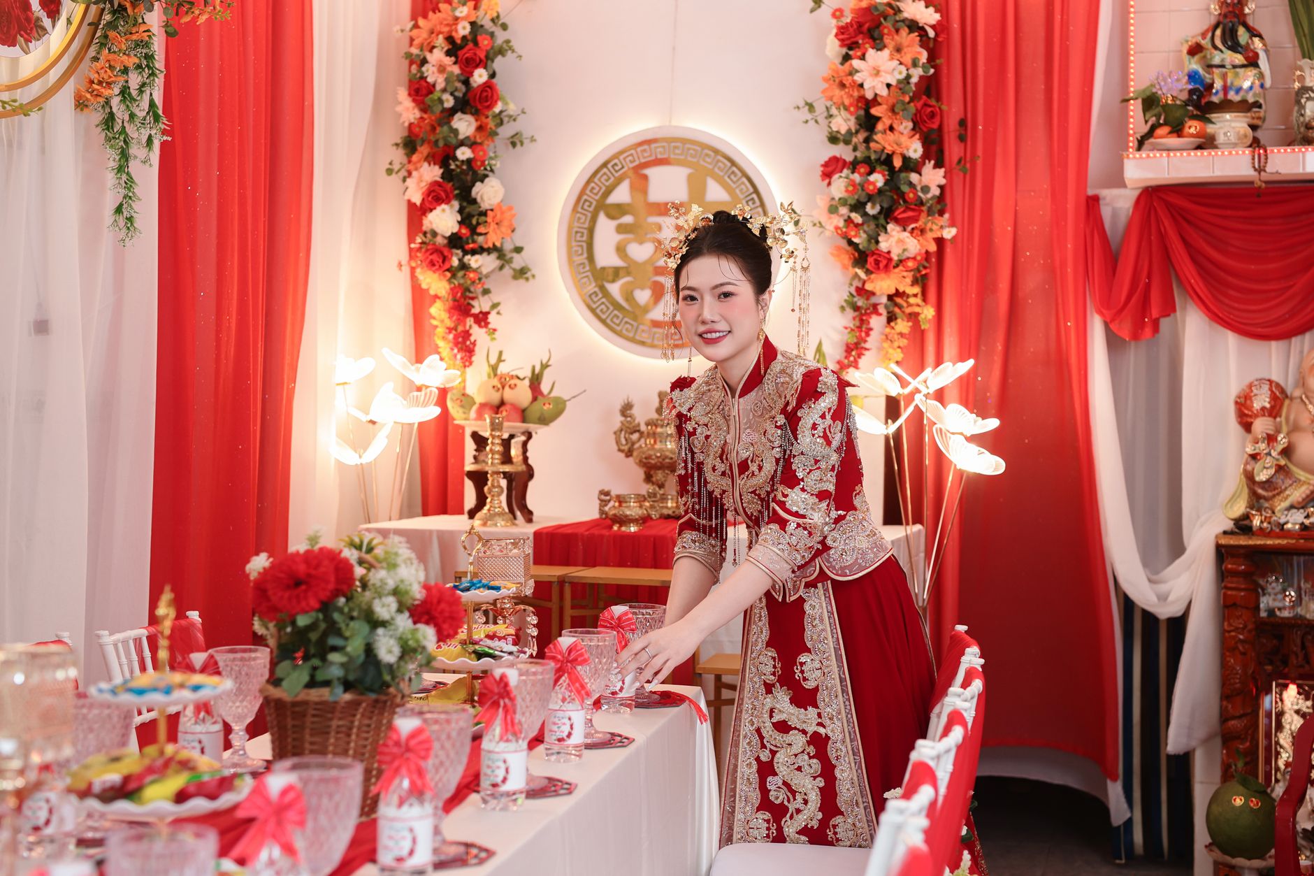A woman in traditional attire sets an elegant table with red floral decorations at an Asian wedding.